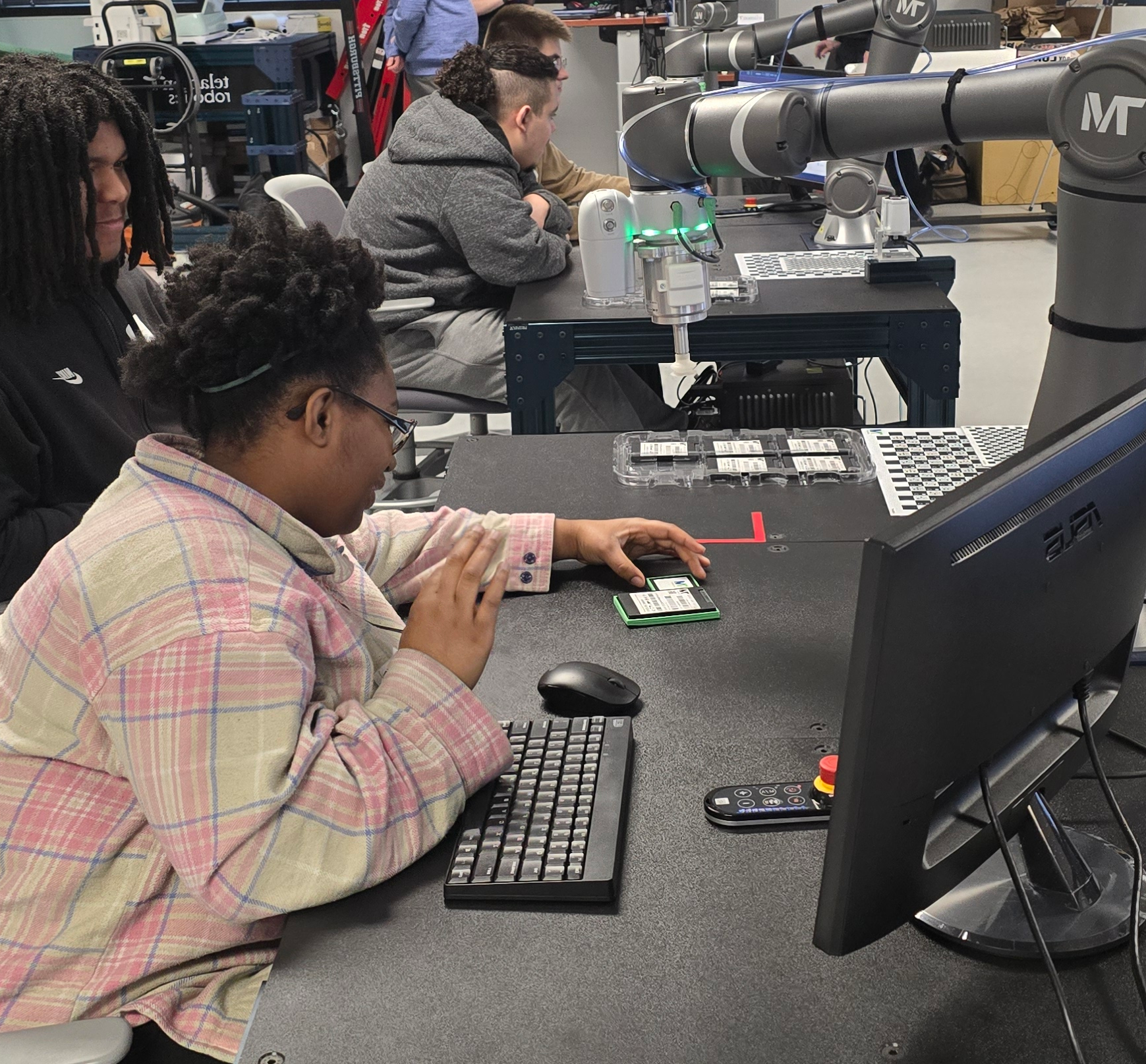 Four high school diverse male and female students sitting in pairs at stations with collaborative robots and computers, interacting with the technology